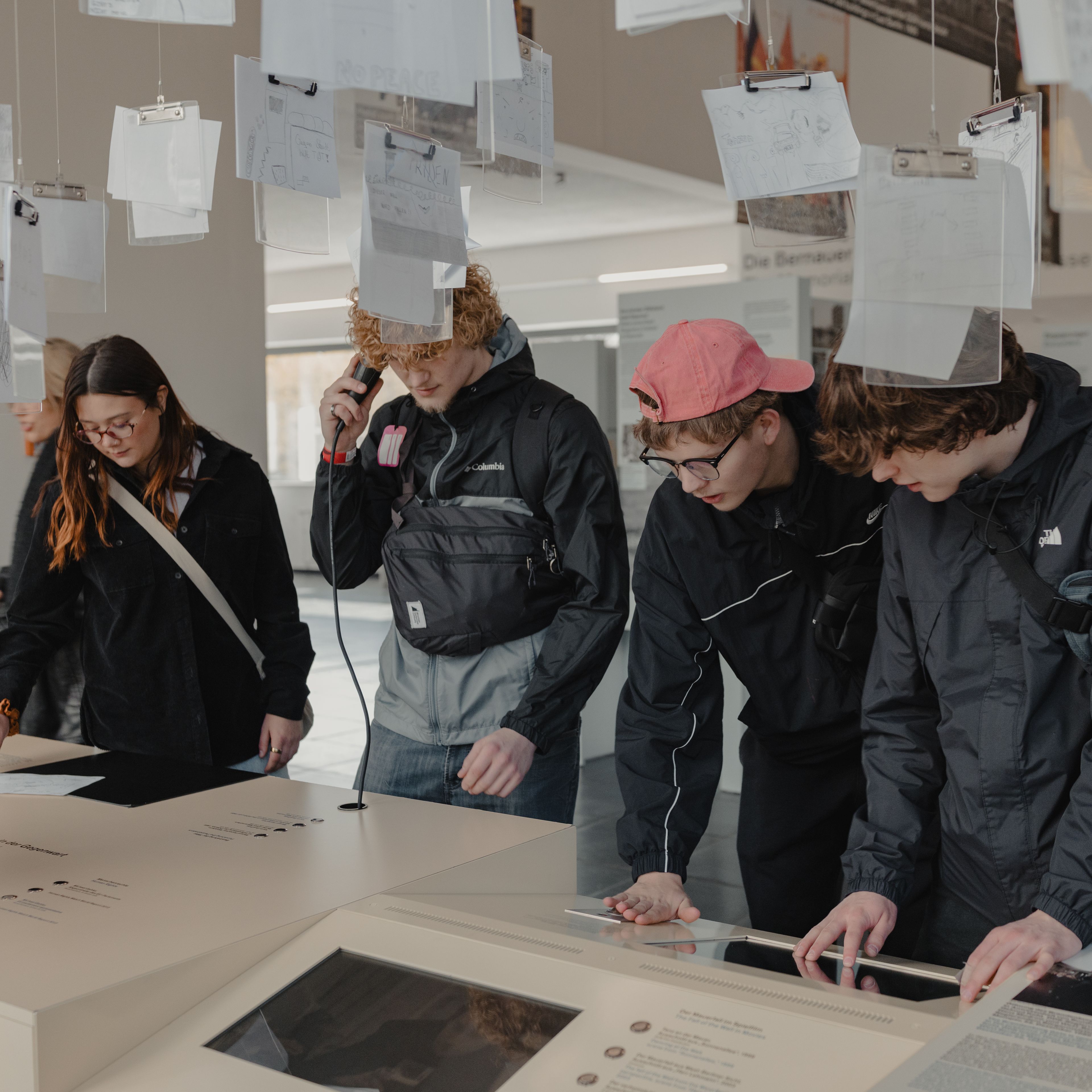 Three students and their teacher leaning over interactive display tables and listening to an audio handset in a Berlin Wall Memorial exhibit, with papers on clipboards hanging overhead