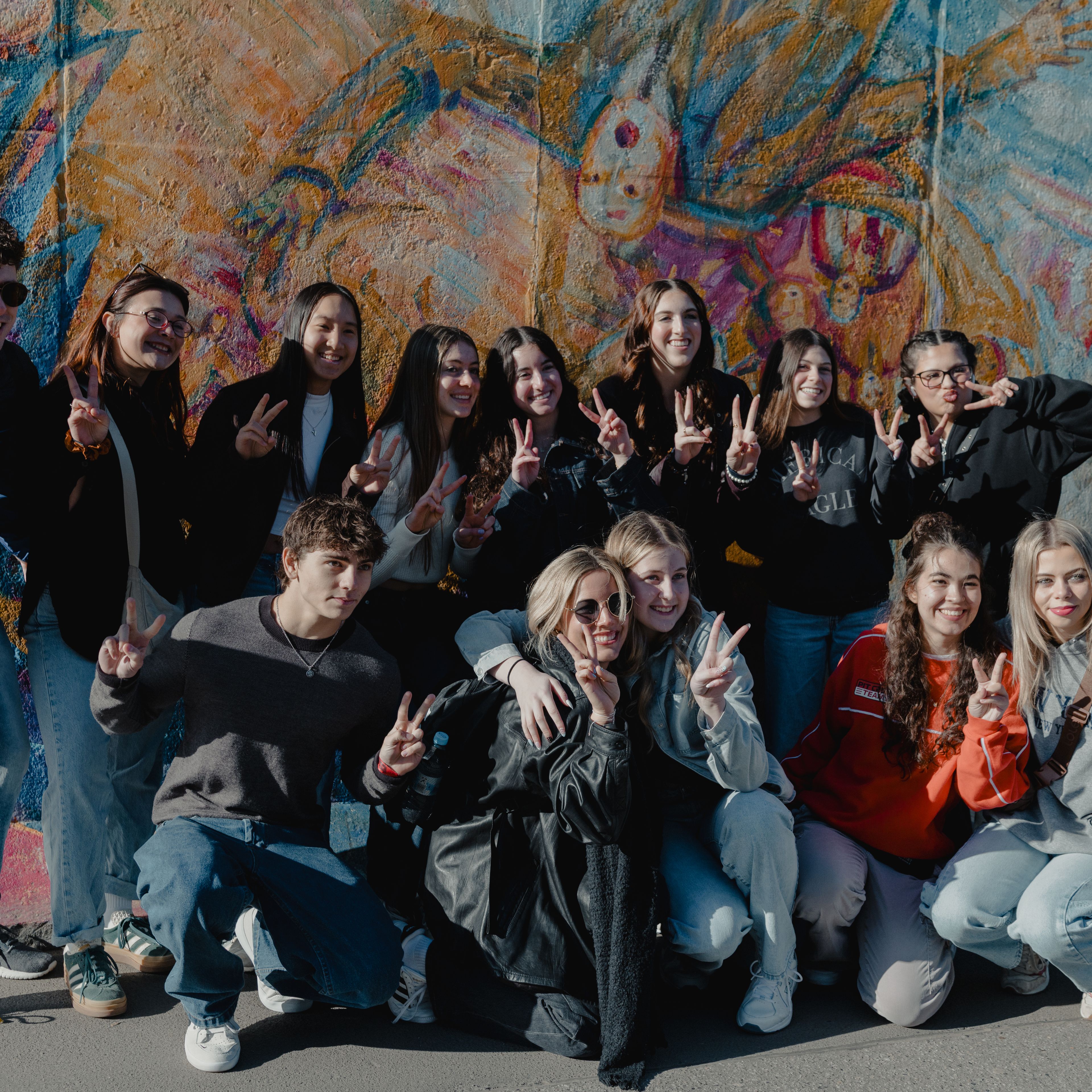 Group of students posing with peace signs, some standing and some crouching, in front of a large colourful painted mural in Germany