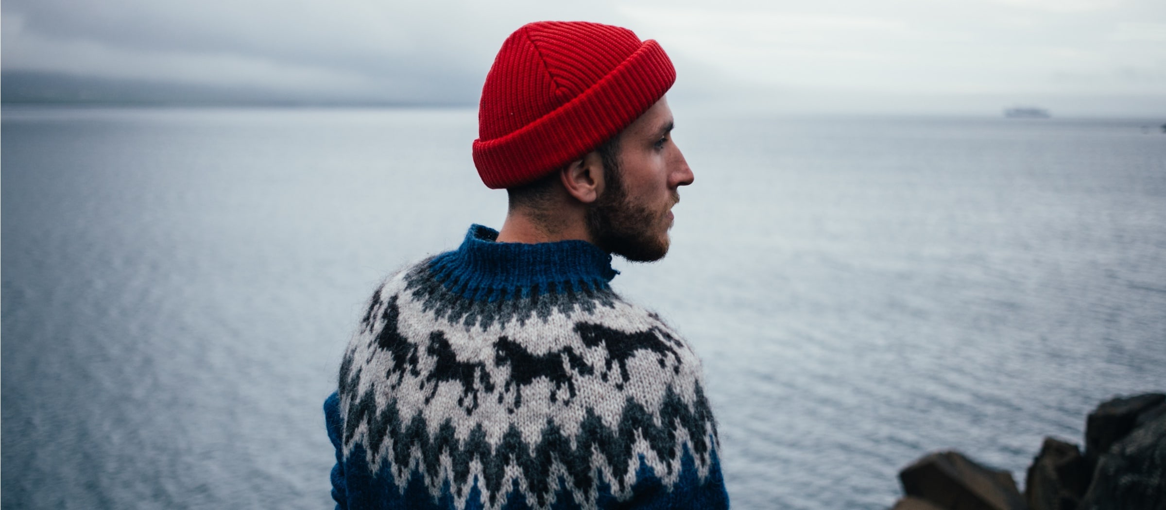 Ein Mann mit roter Mütze und gemustertem Pullover blickt auf das Meer, im Hintergrund sind ein bewölkter Himmel und ein fernes Land zu sehen.
