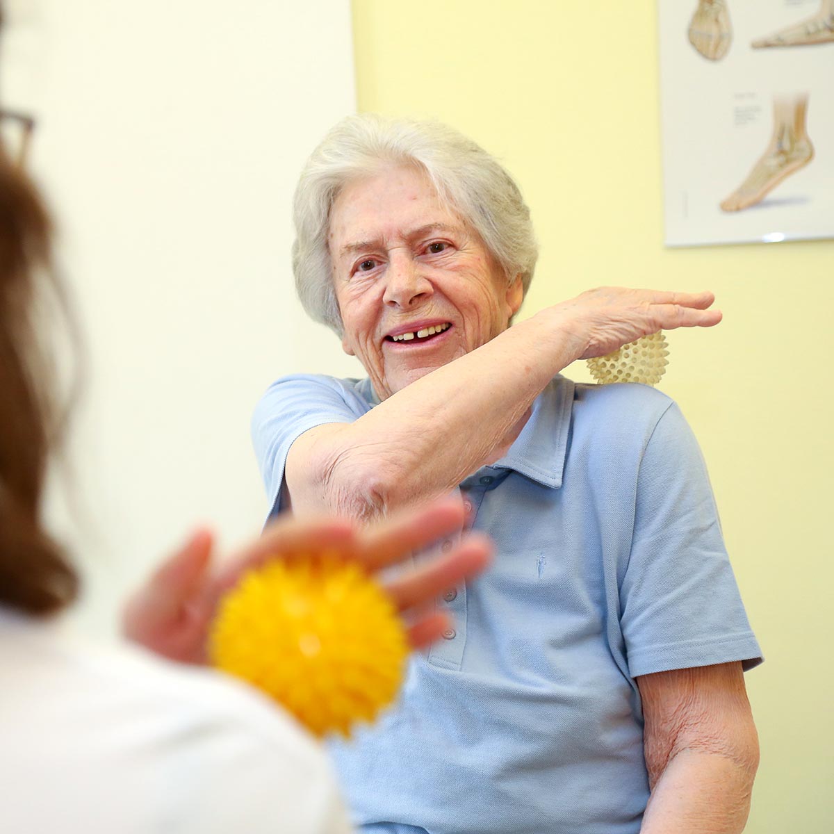 Eine ältere Dame massiert nach Anleitung einer Therapeutin ihre Schulter mit einem Therapieball.