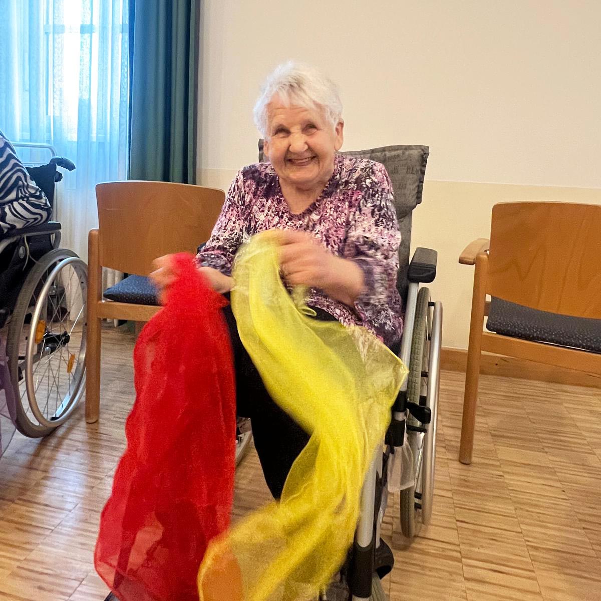 Eine ältere Dame im Rollstuhl tanzt mit zwei bunten Tüchern.