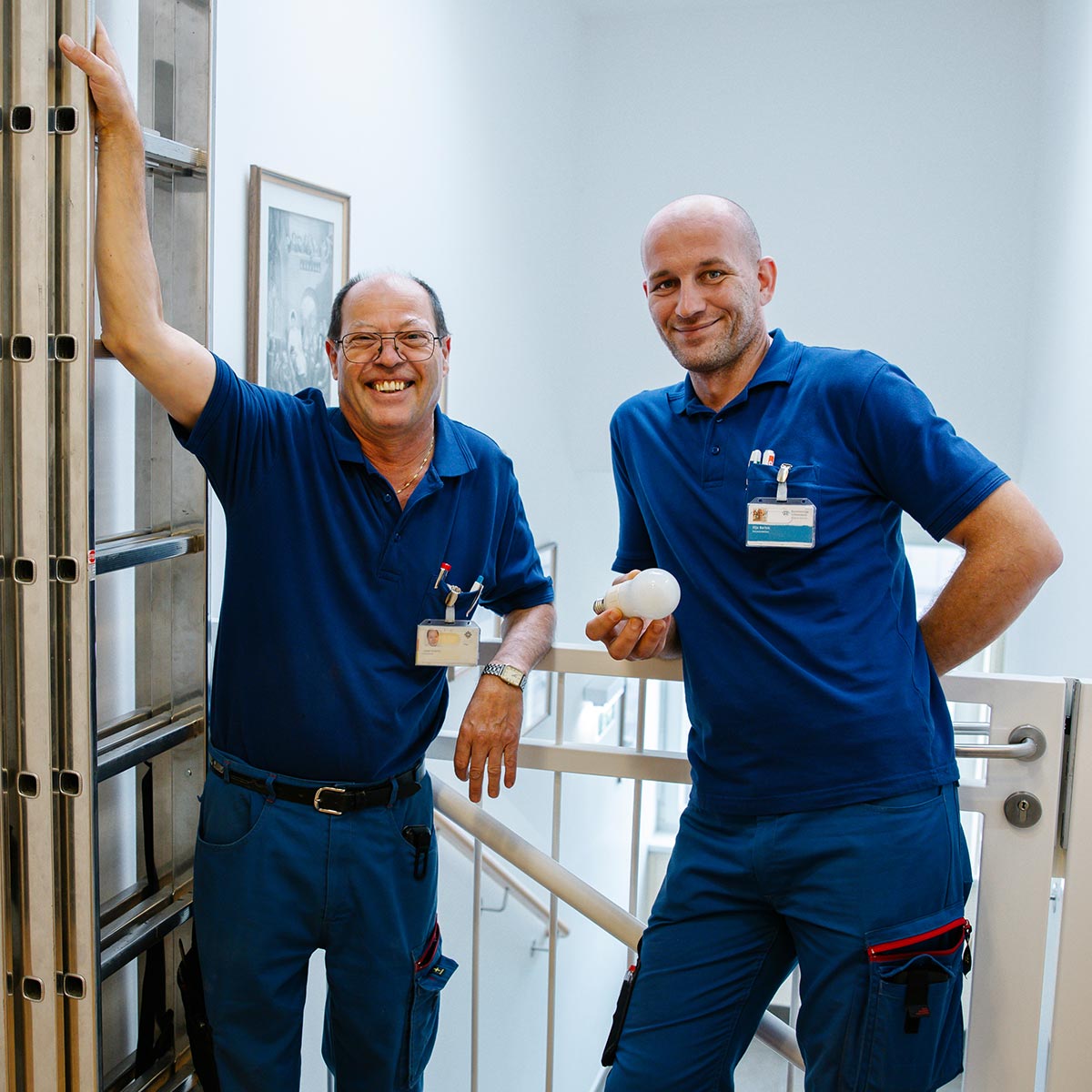 Zwei Haustechniker lehnen gelassen bei der Leiter mit Glühbirne in der Hand