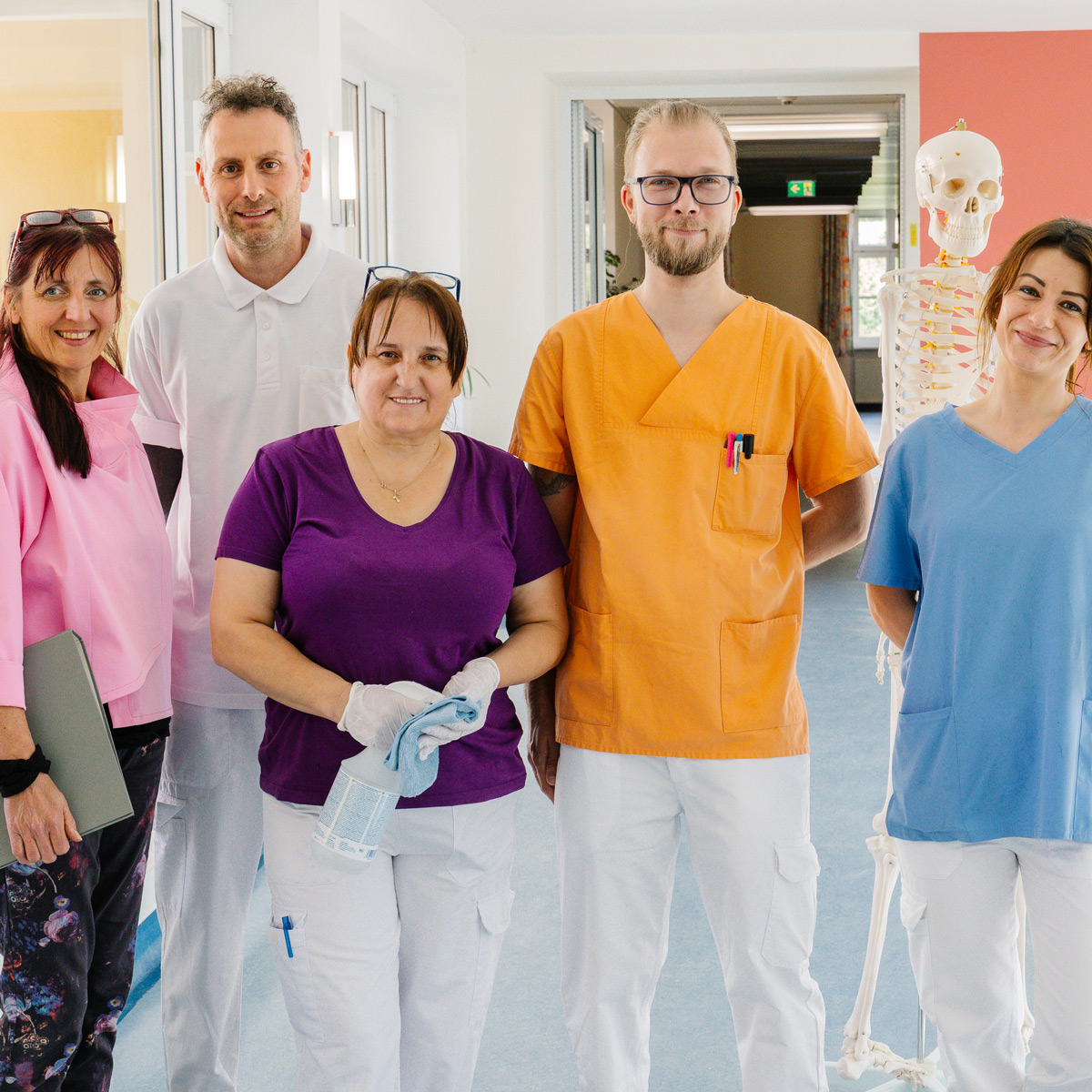Mitarbeiter*innen aus Verwaltung, Pflege, Therapie und Reinigung stehen in Dienstkleidung lächelnd nebeneinander