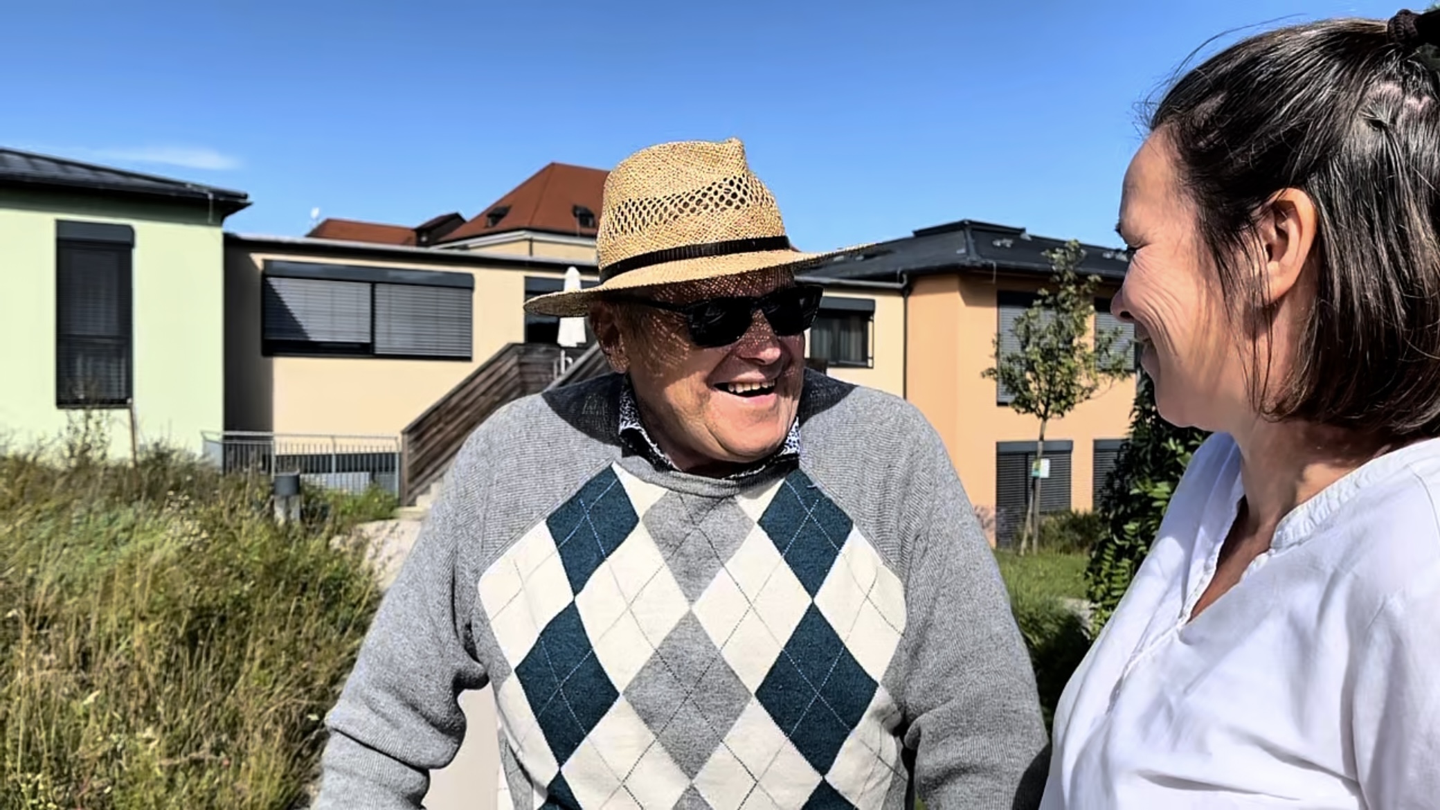 Ein Bewohner mit Sonnenbrille und Hut unterhält sich mit einer Mitarbeiterin im Garten des Pflegehauses St. Louise.
