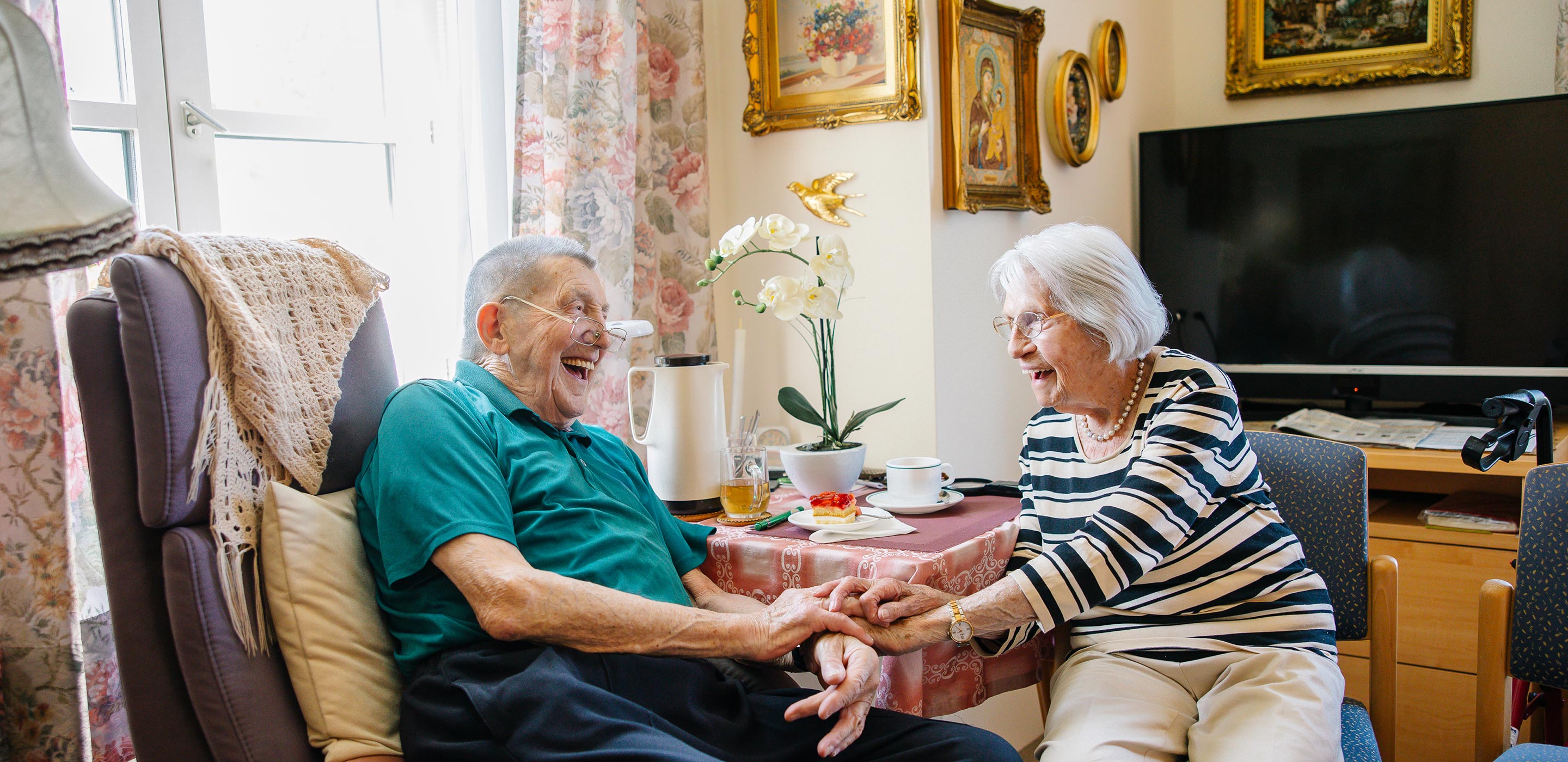 Ein älterer Mann und eine ältere Frau halten die Hände und lächeln einander freundschaftlich an. Sie sitzen bem Tisch in der gemütlich eingerichteten Wohnung.