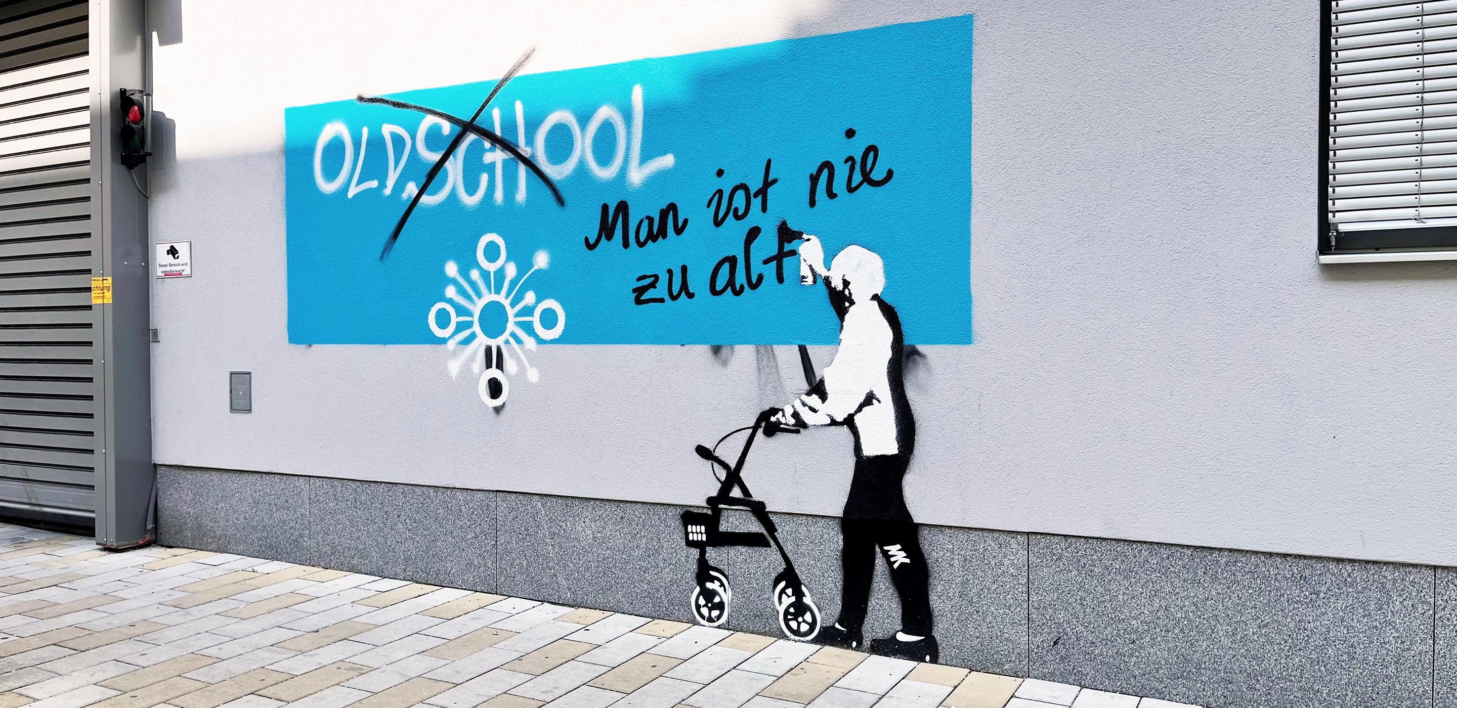 Ein Graffiti auf der Hausmauer zeigt eine ältere Dame mit Rollator, die mit Sprüdose schreibt: "Oldschool, durchgestrichen. Man ist nie zu alt!"