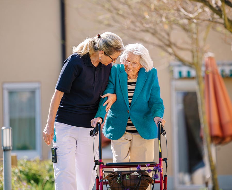 Eine Betreuerin geht gemeinsam mit einer älteren Dame mit Rollator im Garten spazieren