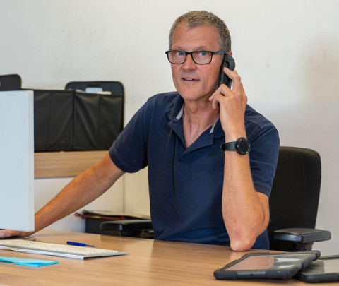 Man met bril en horloge, zittend aan een bureau, bellend, met een computer en tablet in de buurt.
