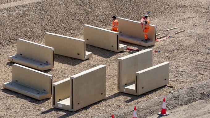 Collection of concrete Jersey barriers and other precast concrete elements are neatly organized on a gravel surface at a construction site, awaiting installation