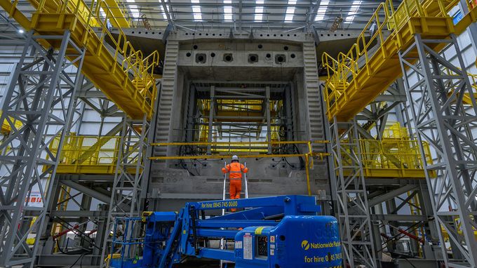 Interior of first deck segment in factory with blue scissor lift in foreground and worker in orange safety gear.