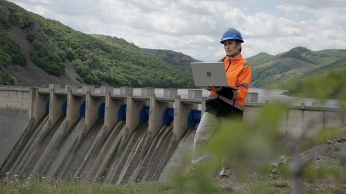 Person in safety gear using a laptop near a large dam.