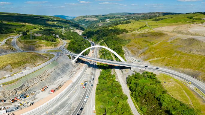 Aerial view of a highway bridge under construction in a lush valley, with vehicles on roads, green fields, and blue sky with clouds in the background.