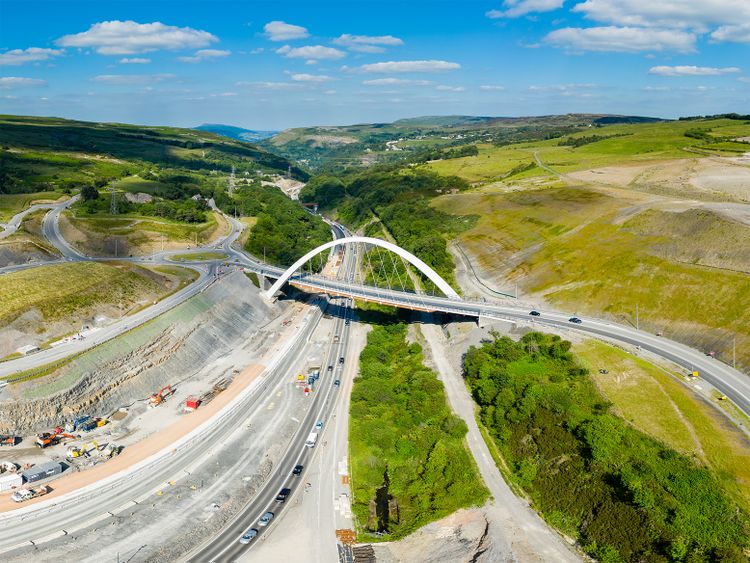 Aerial view of a highway bridge under construction in a lush valley, with vehicles on roads, green fields, and blue sky with clouds in the background.