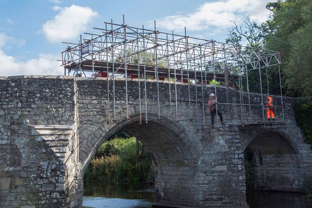 Damaged bridge over a river with scaffolding on top and two workers in safety gear inspecting the structure.