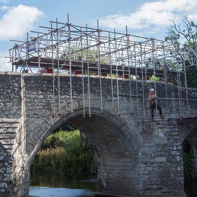 Damaged bridge over a river with scaffolding on top and two workers in safety gear inspecting the structure.