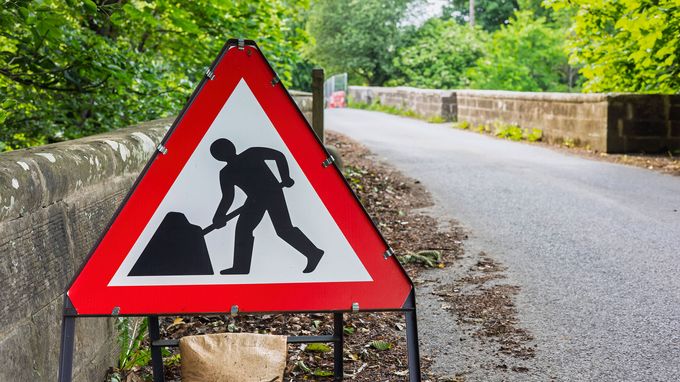 Triangular roadwork sign with a person digging, placed on a rural road lined with greenery and a stone wall.