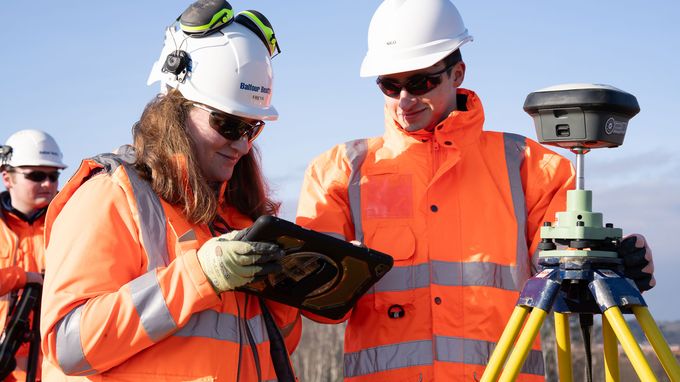 Two construction surveyors in orange high-vis jackets and hard hats use a tablet and tripod-mounted GNSS receiver