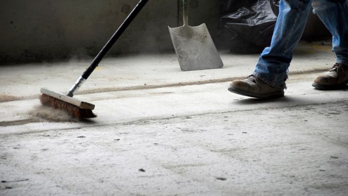 A person in jeans and boots sweeping a concrete floor with a broom, a shovel leaning nearby, and dust visible in the air.