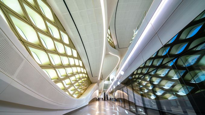 Futuristic hallway with curved, illuminated ceiling and wall patterns, mirrored reflections on right. People visible in the distance.