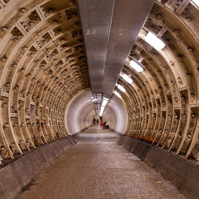 Dimly lit underground tunnel with exposed beams, showing a curved, ribbed structure. Two people are visible in the distance.