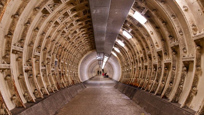Dimly lit underground tunnel with exposed beams, showing a curved, ribbed structure. Two people are visible in the distance.