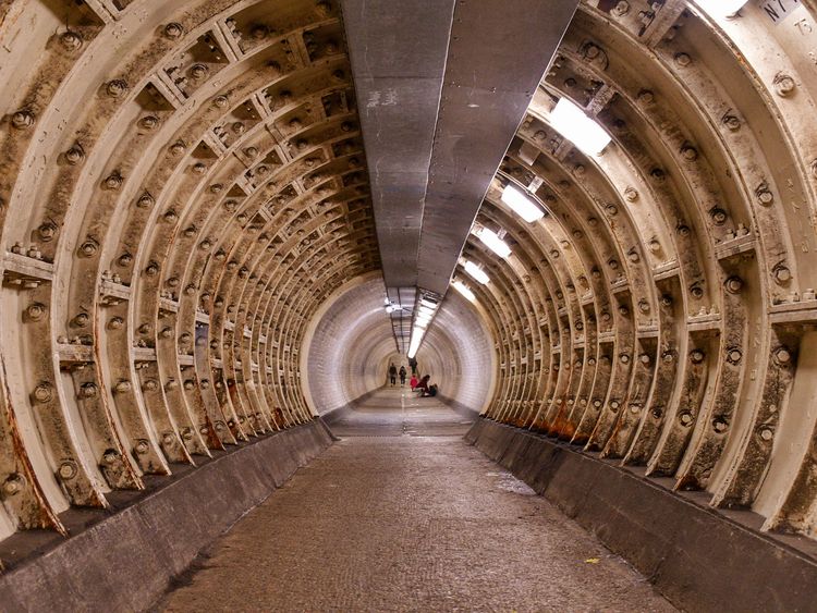 Dimly lit underground tunnel with exposed beams, showing a curved, ribbed structure. Two people are visible in the distance.