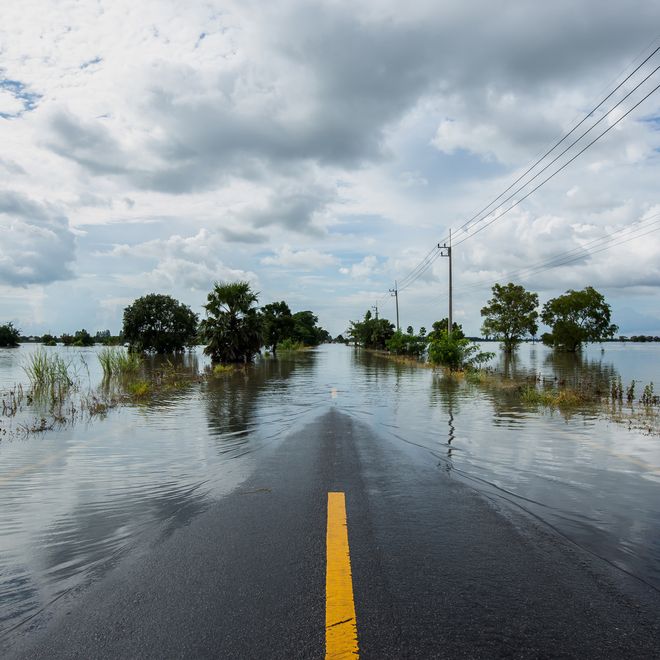 Flooded road with a visible yellow line, surrounded by water and sparse trees under a cloudy sky. Power lines run alongside the road.