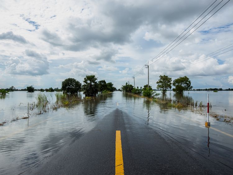 Flooded road with a visible yellow line, surrounded by water and sparse trees under a cloudy sky. Power lines run alongside the road.