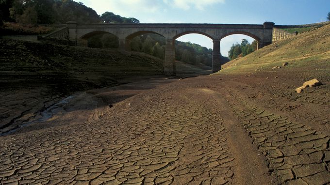 Dry riverbed with cracked earth beneath an old stone bridge, surrounded by grassy slopes and trees under a partly cloudy sky.