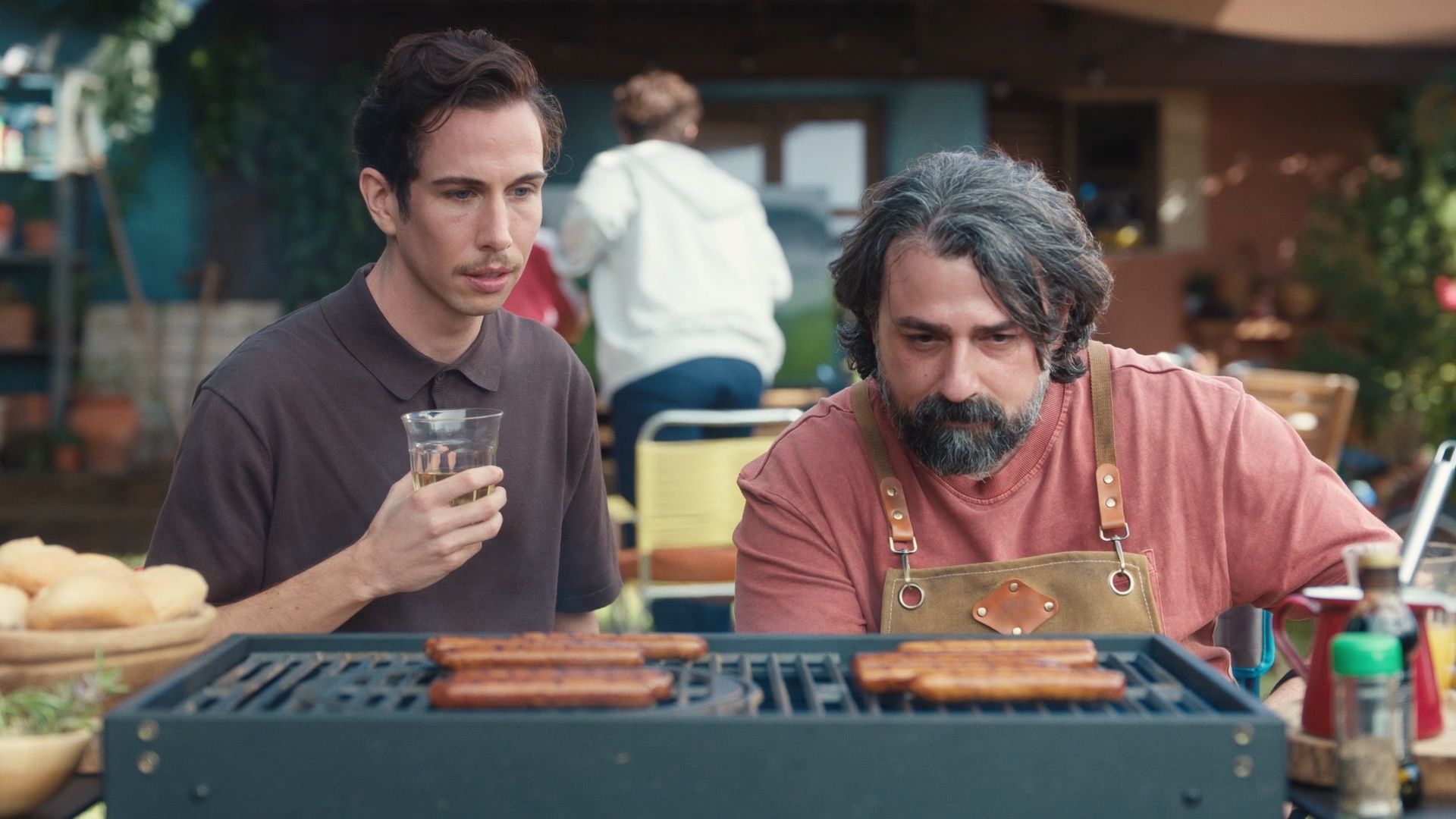 Two men focused on grilling sausages outdoors. One holds a drink, the other wears an apron. Bread and condiments are visible nearby.