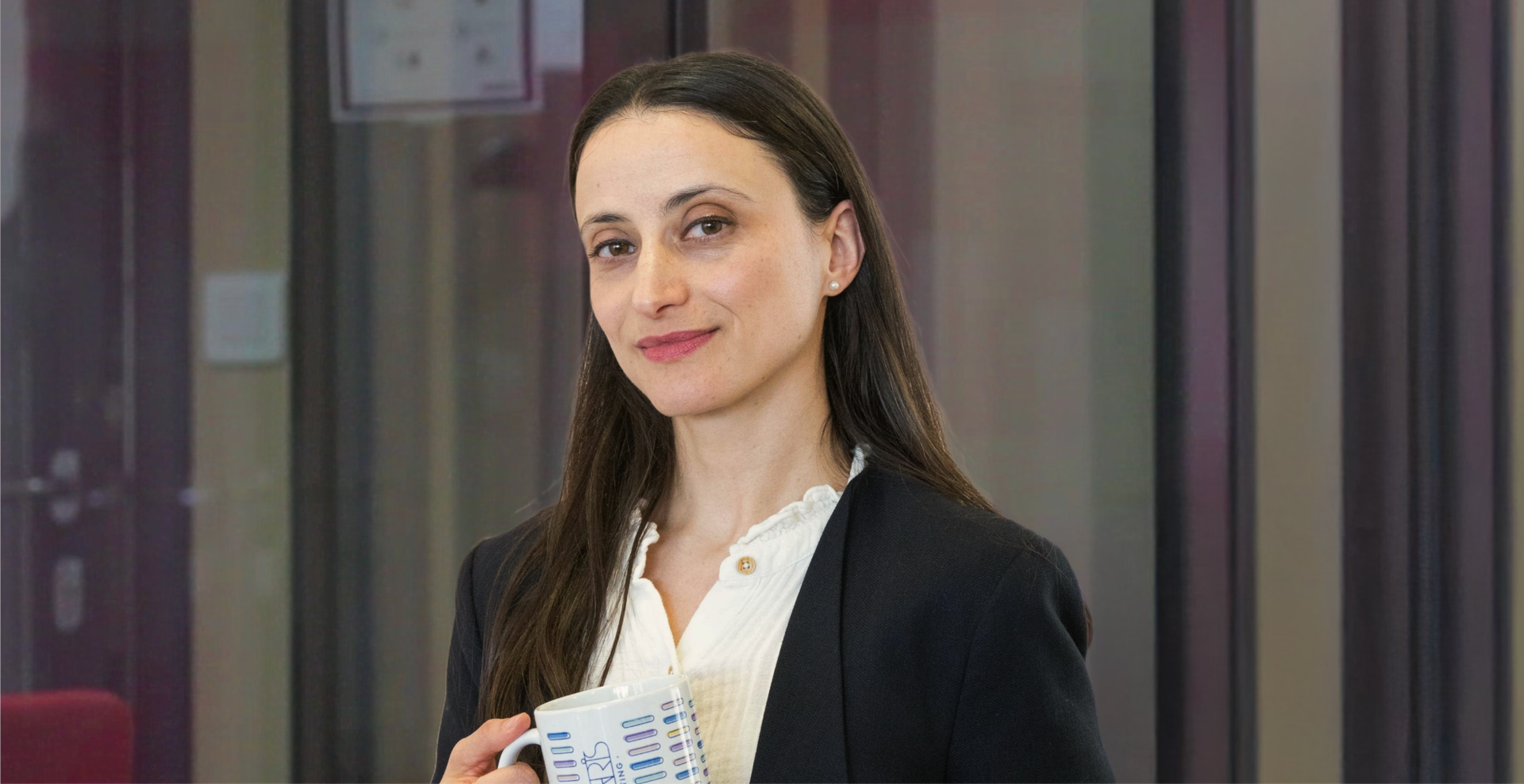 Woman in a blazer holding a mug, standing confidently in an office setting.
