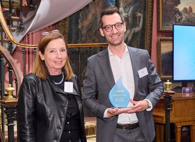 Man in suit holding blue teardrop award beside woman in leather jacket, posed in ornate interior with staircase and paintings.