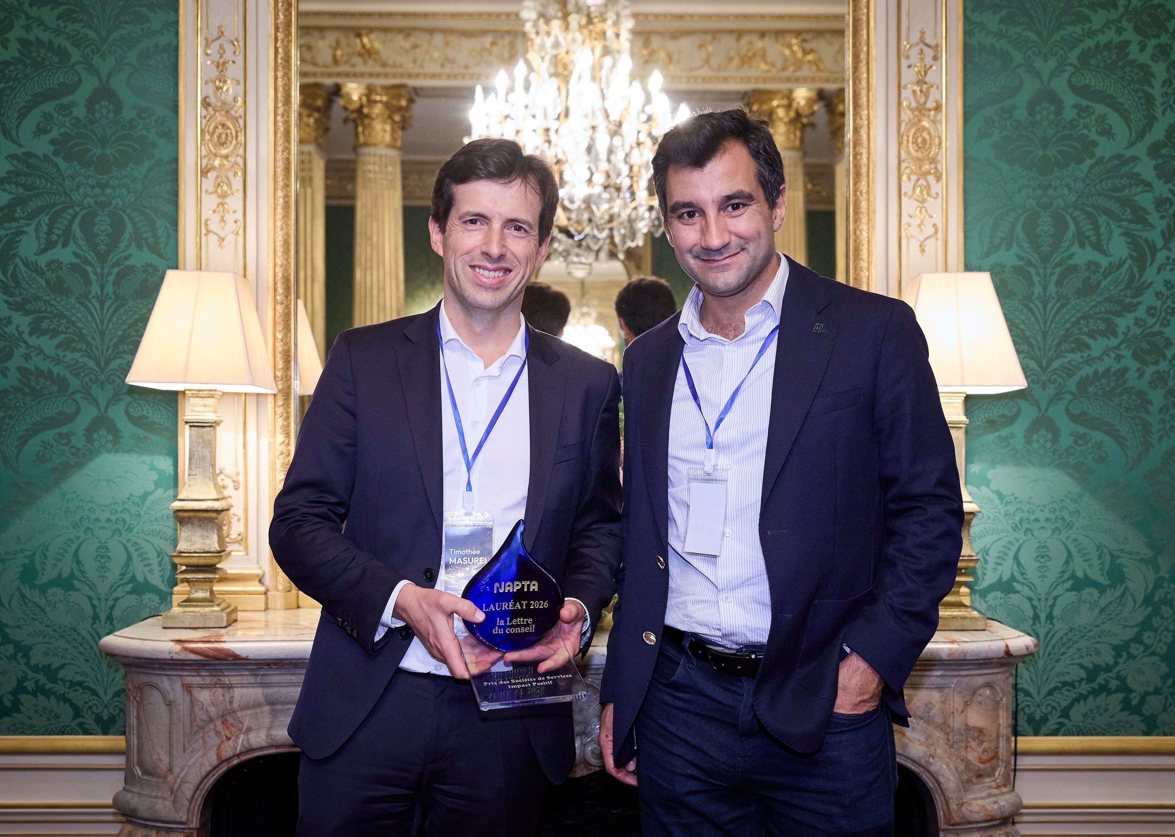 Two men in suits smiling, one holding a blue award, standing in an elegant room with a chandelier and ornate decor.
