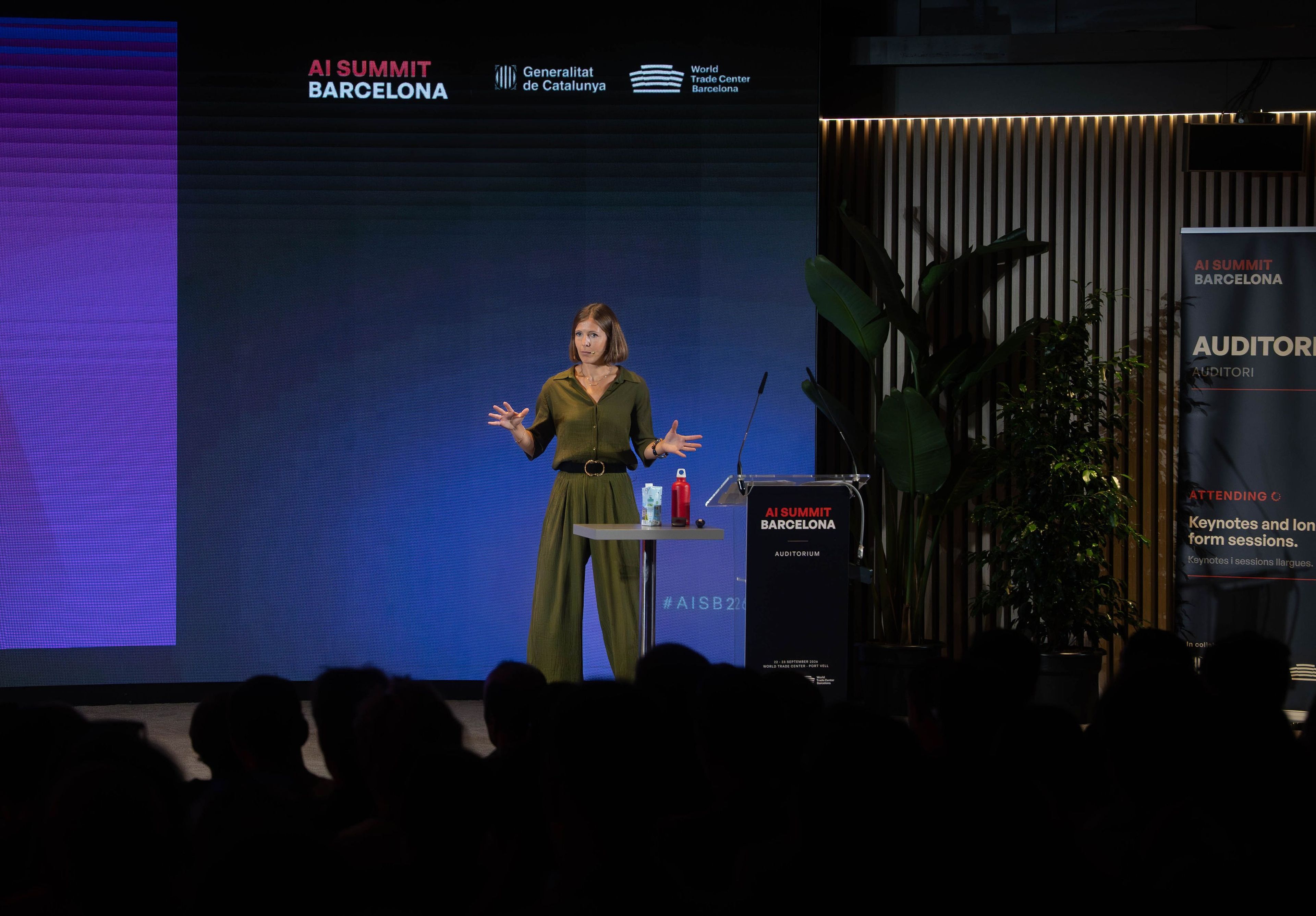 A woman speaks on stage at the AI Summit Barcelona, standing beside a podium with a microphone, in front of a screen displaying event branding.
