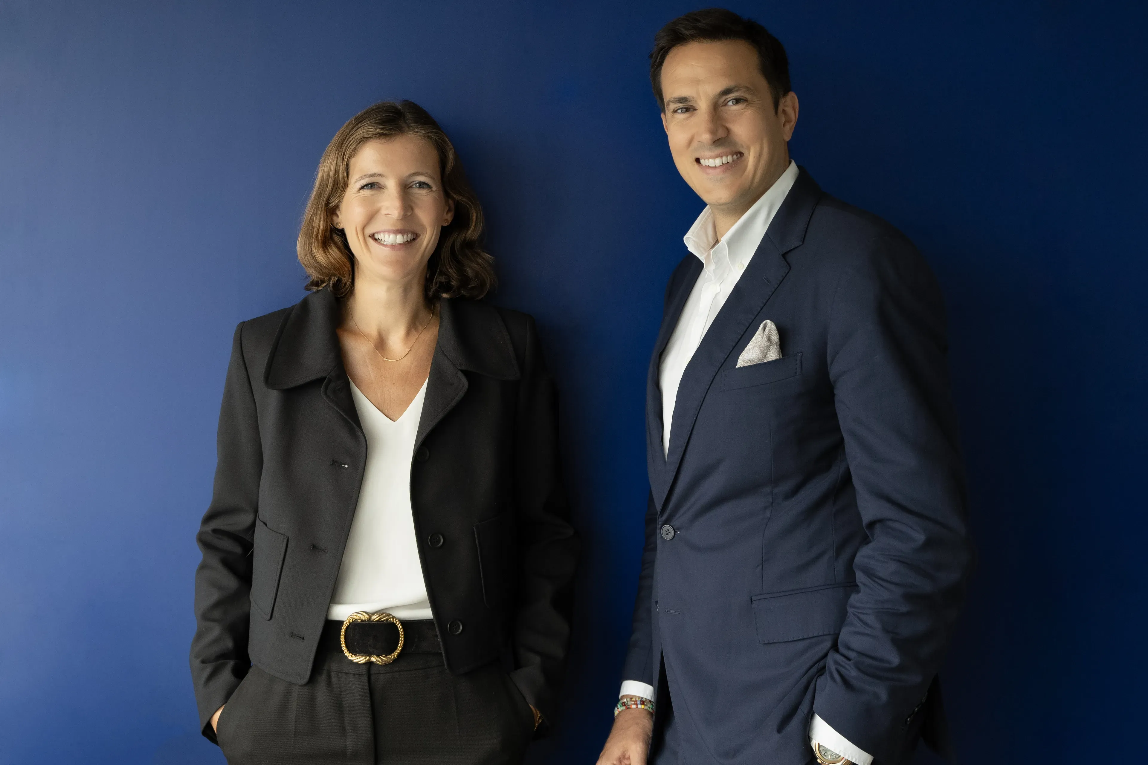 Two people smiling, standing against a blue background. The woman wears a black jacket, and the man wears a navy suit with a white shirt.