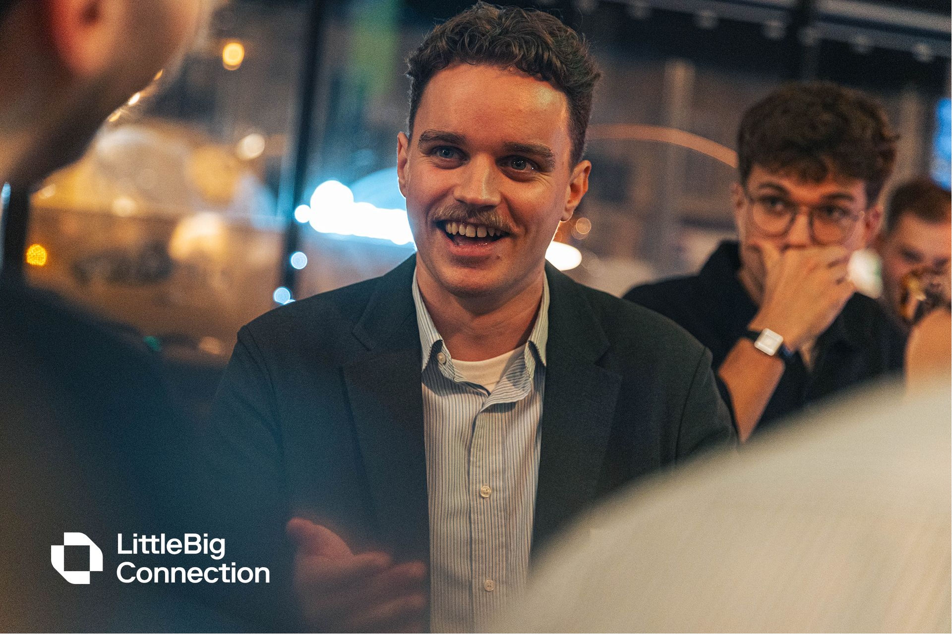 A man with a mustache smiles and talks animatedly at an indoor gathering. A person in the background listens intently.