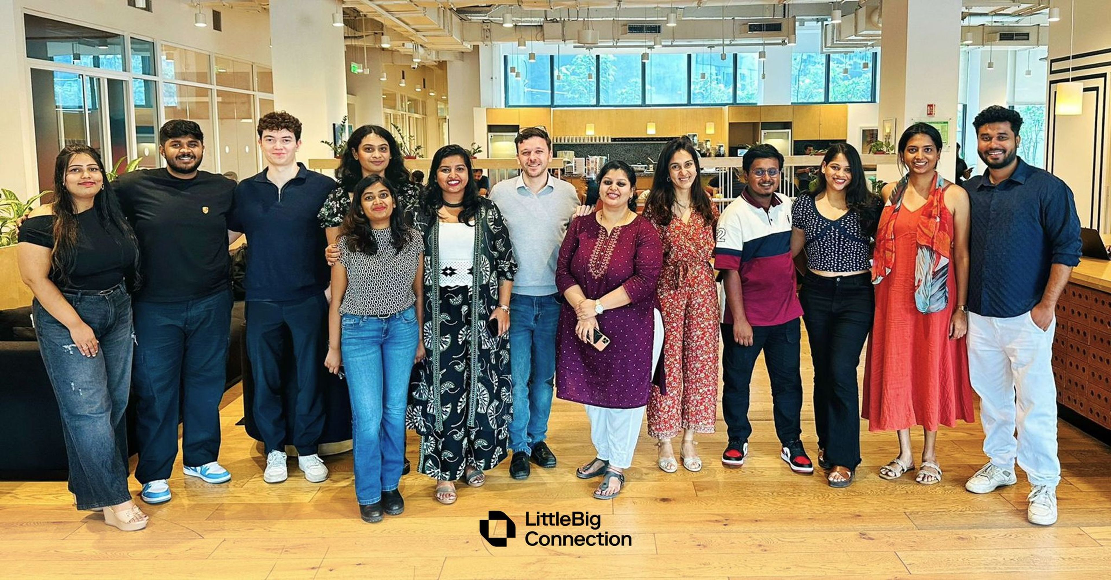 A group of diverse people standing together in an office environment, smiling. The room has large windows and a modern interior design.