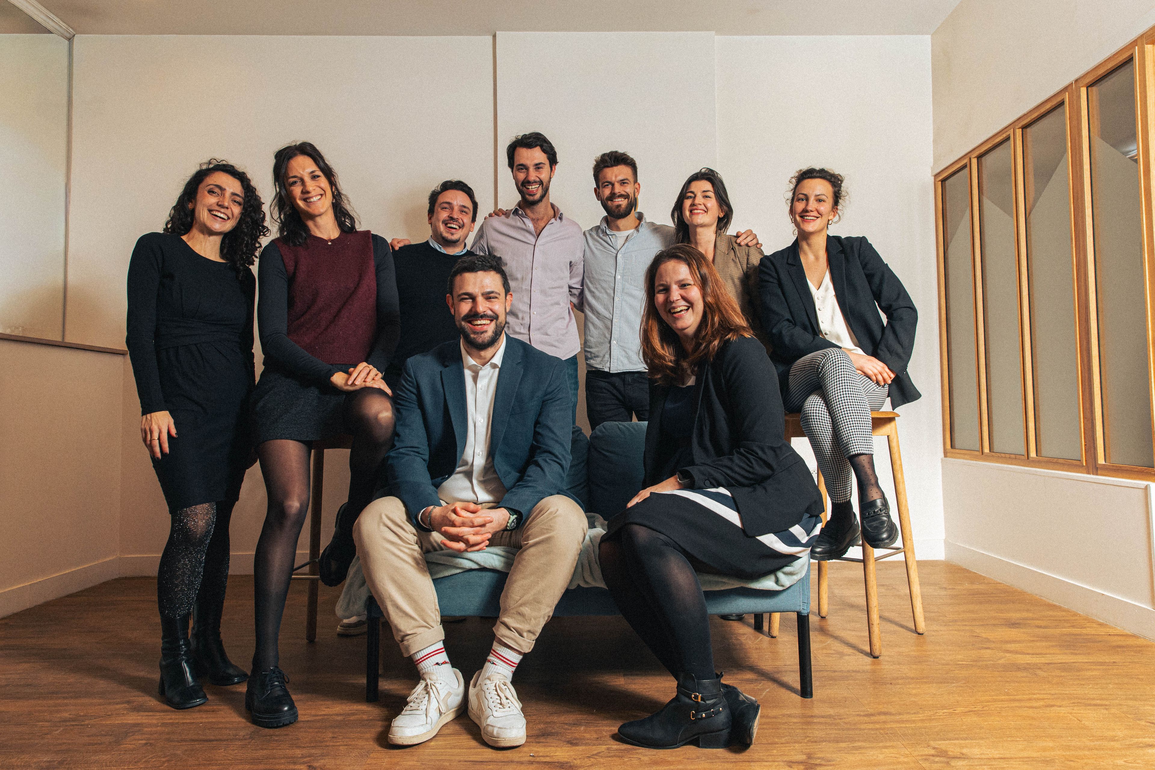 A group of nine people posing for a photo indoors, smiling and casually dressed, in front of a neutral background.