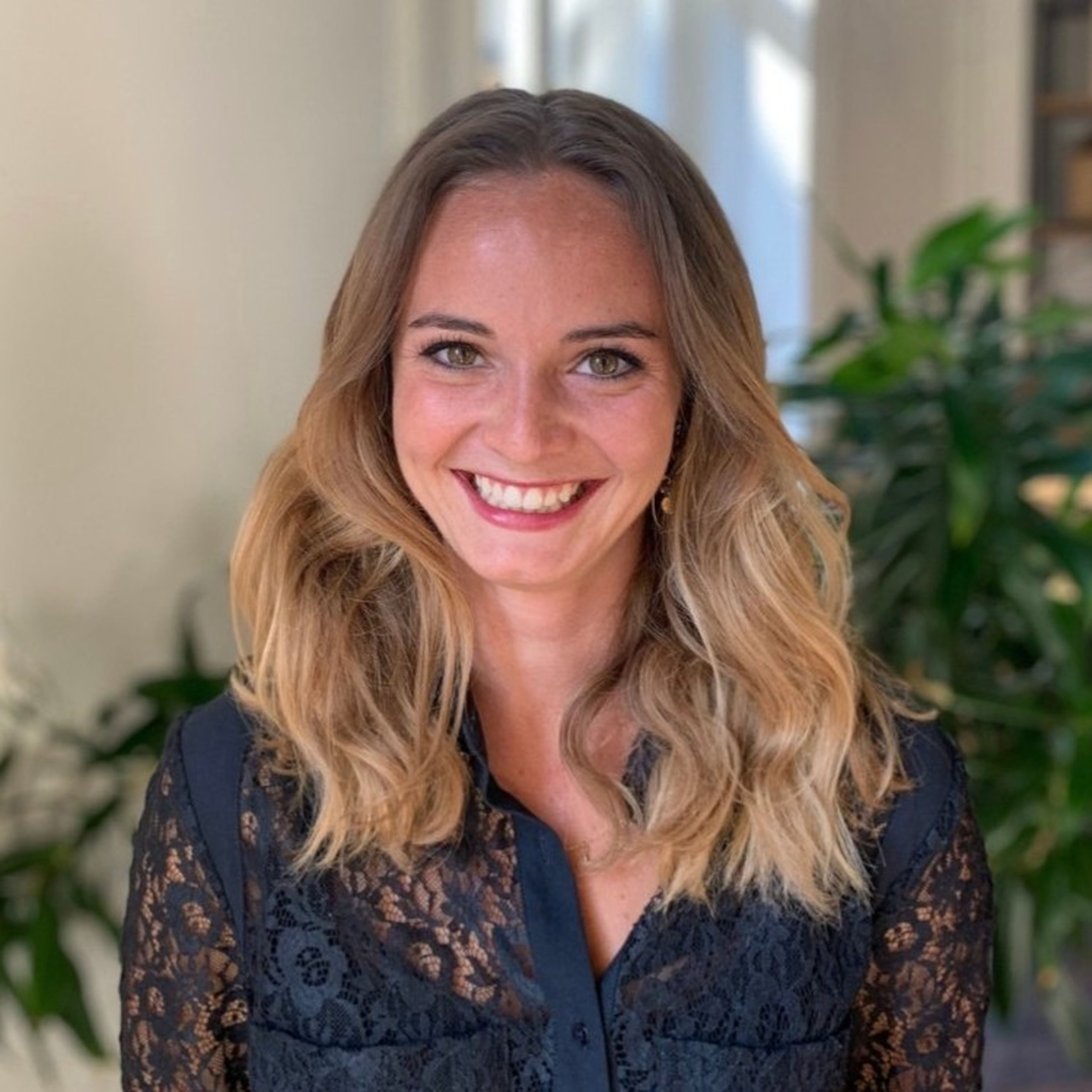 Smiling woman with long wavy hair, wearing a black lace top, standing in front of a plant indoors.