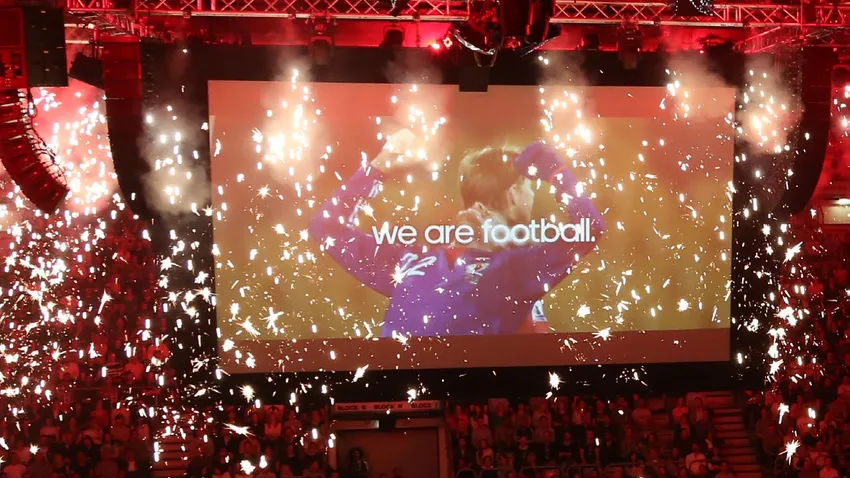Large screen with "we are football" text, surrounded by bright sparkles and red lighting, at a packed indoor event.