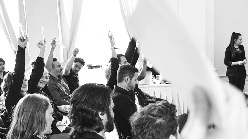 Audience members in a meeting raise cards to vote, while a woman stands and observes. The scene is in black and white.