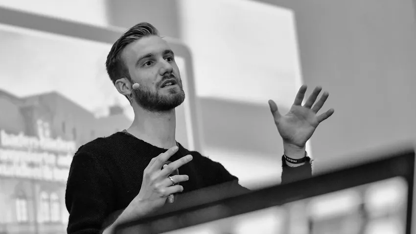 A person speaking energetically on stage with a headset microphone, using expressive hand gestures. Black and white image.