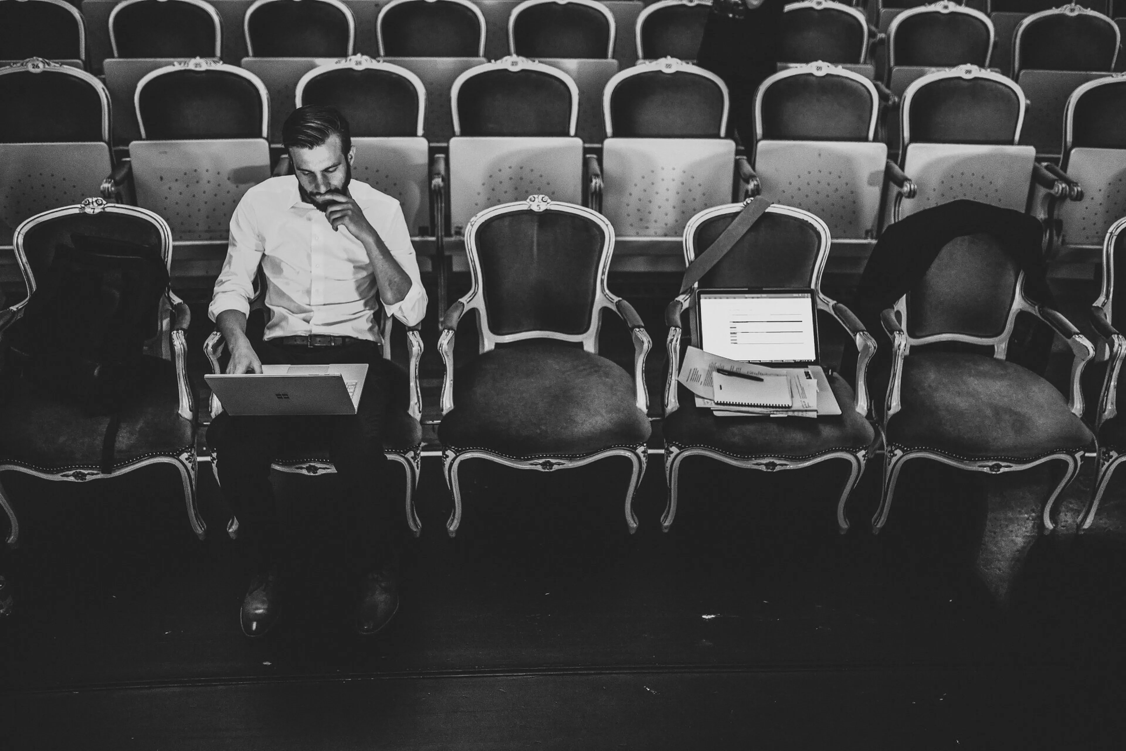 A person sits in an empty theater with ornate chairs, working on a laptop, accompanied by open documents on an adjacent seat. Black and white image.