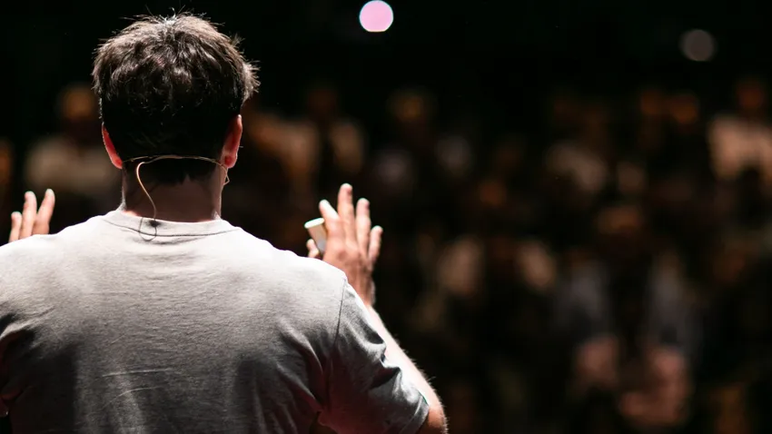 Back view of a person speaking on stage to a blurred audience, wearing a headset microphone, with a dramatic lighting focus.