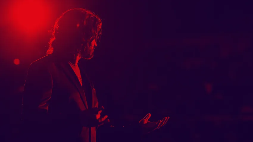 Silhouetted person with long hair and suit gestures on stage under red lighting, creating a dramatic effect.
