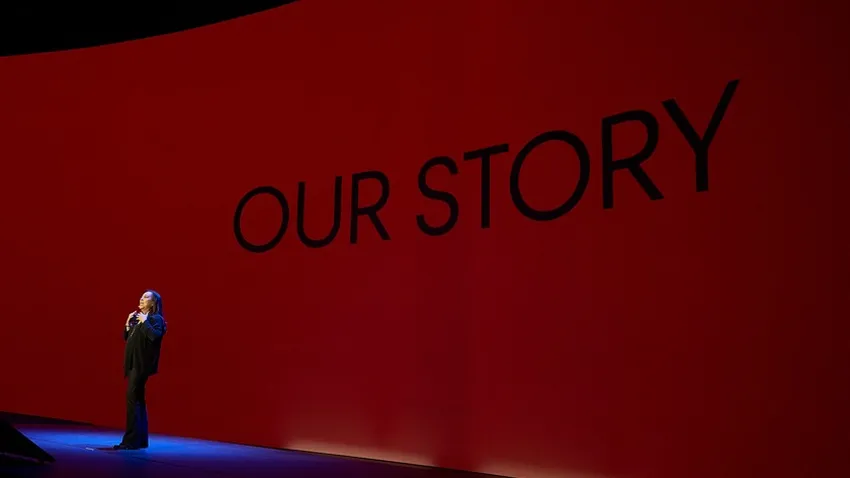 A person stands onstage in front of a red backdrop with "OUR STORY" written in large black letters.
