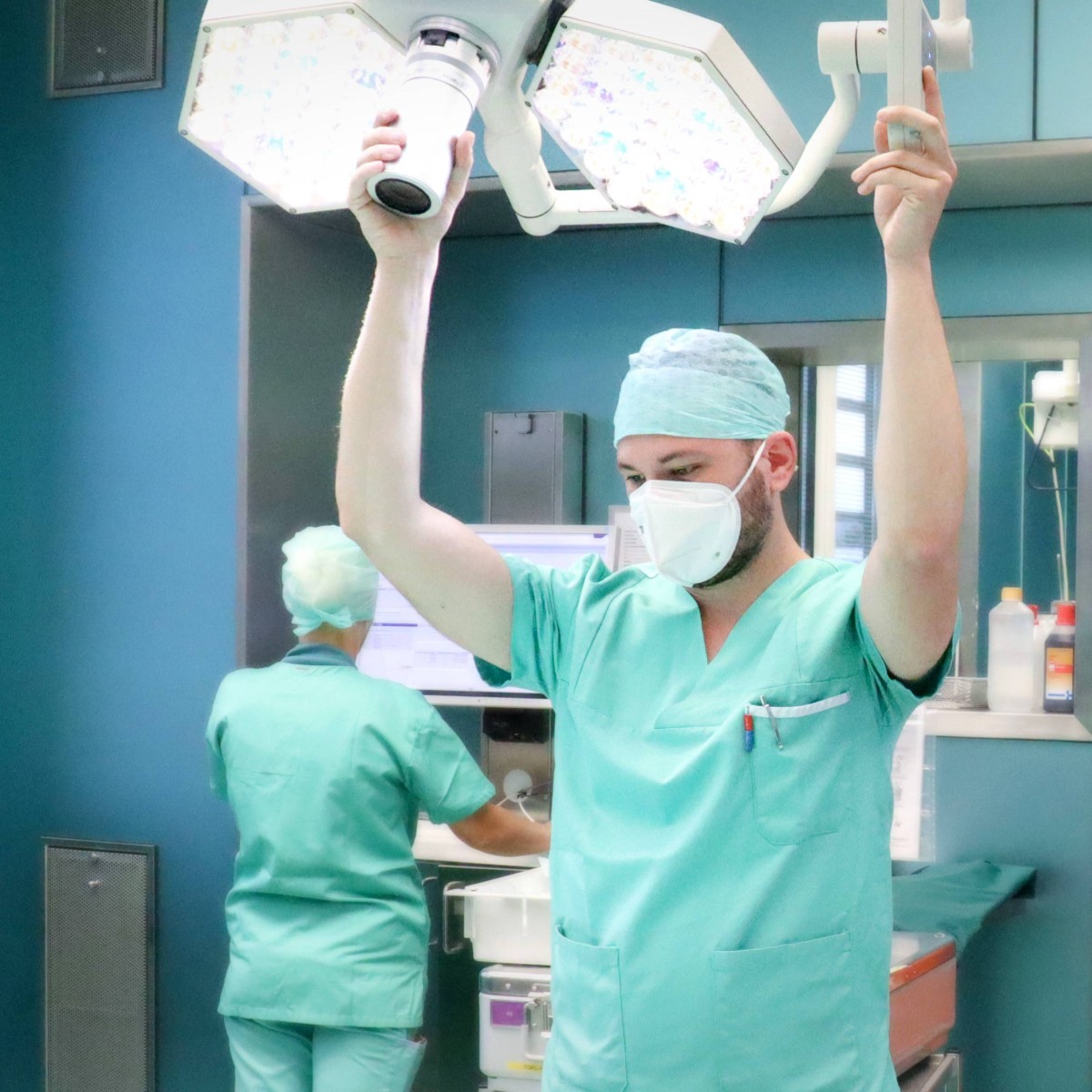 Ein junger Mann in OP-Schutzkleidung und mit Mundmaske steht im OP, er hält mit beiden Händen eine OP-Leuchte und beobachtet das Geschehen.
