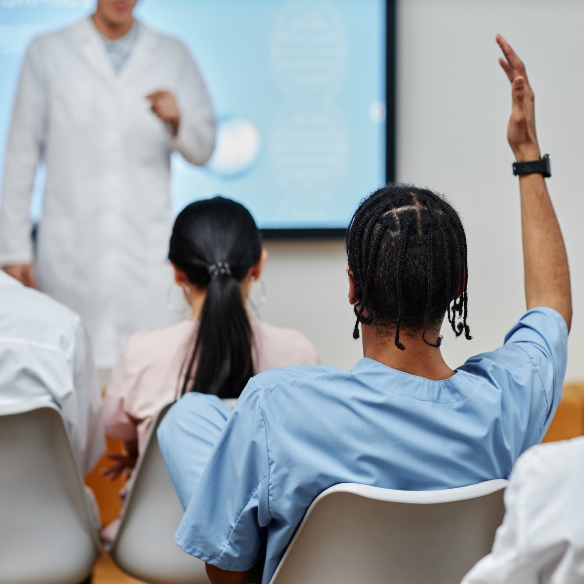 Medizinstudierende in einem Schulungsraum. Vorne steht ein Referent, die Studierenden sind von hinten zu sehen. Einer der Studierenden zeigt auf.