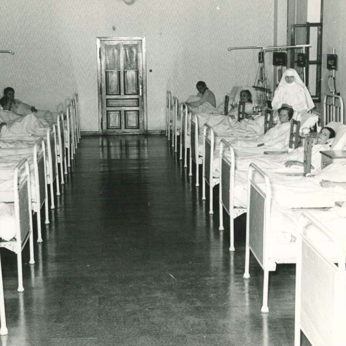 Ein historisches Schwarz-Weiß-Foto. In einer Krankenstation liegen mehrere Männer, zwei Ordensschwestern betreuen sie.