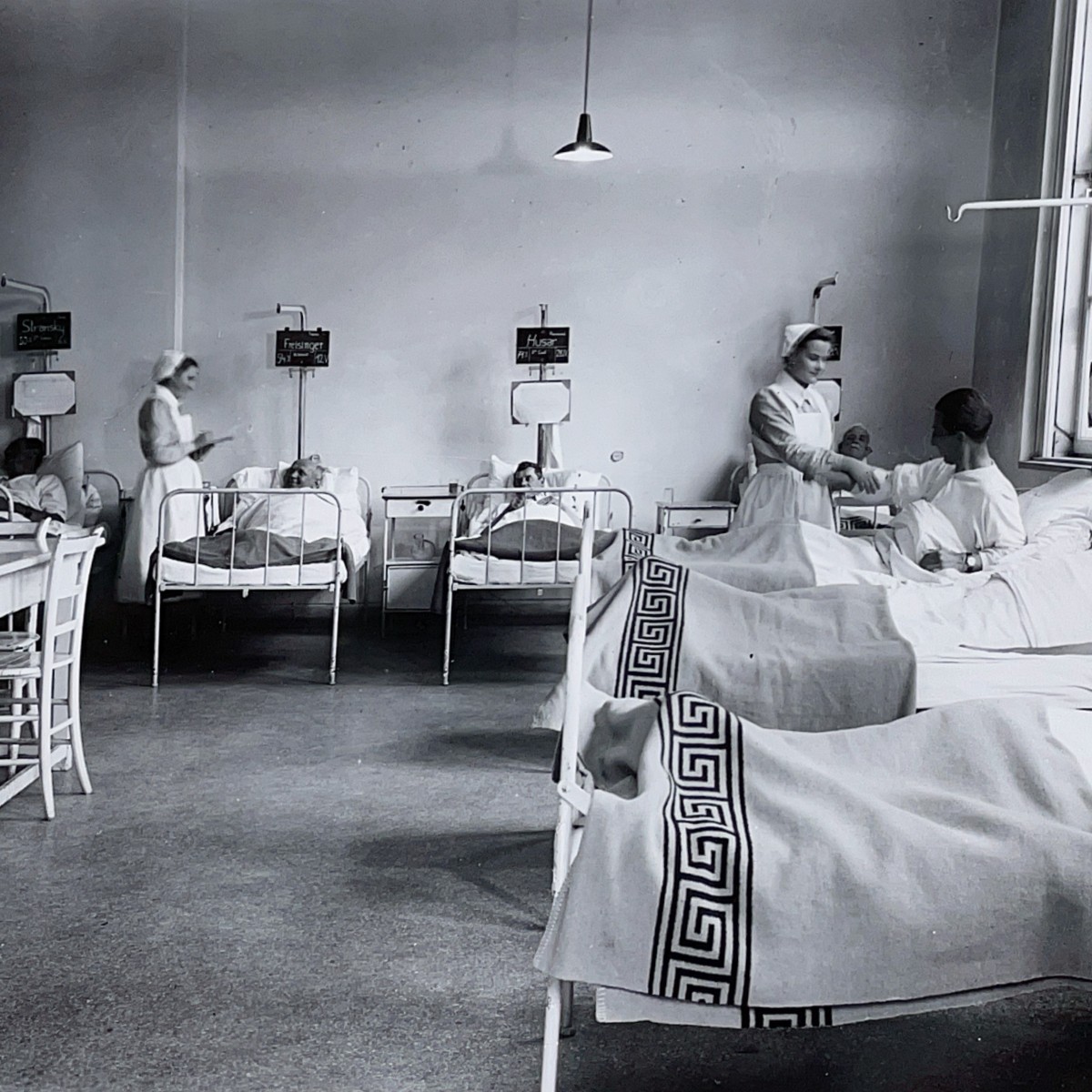 Ein historisches Schwarz-Weiß-Foto. In einer Krankenstation liegen mehrere Männer, zwei Ordensschwestern betreuen sie.